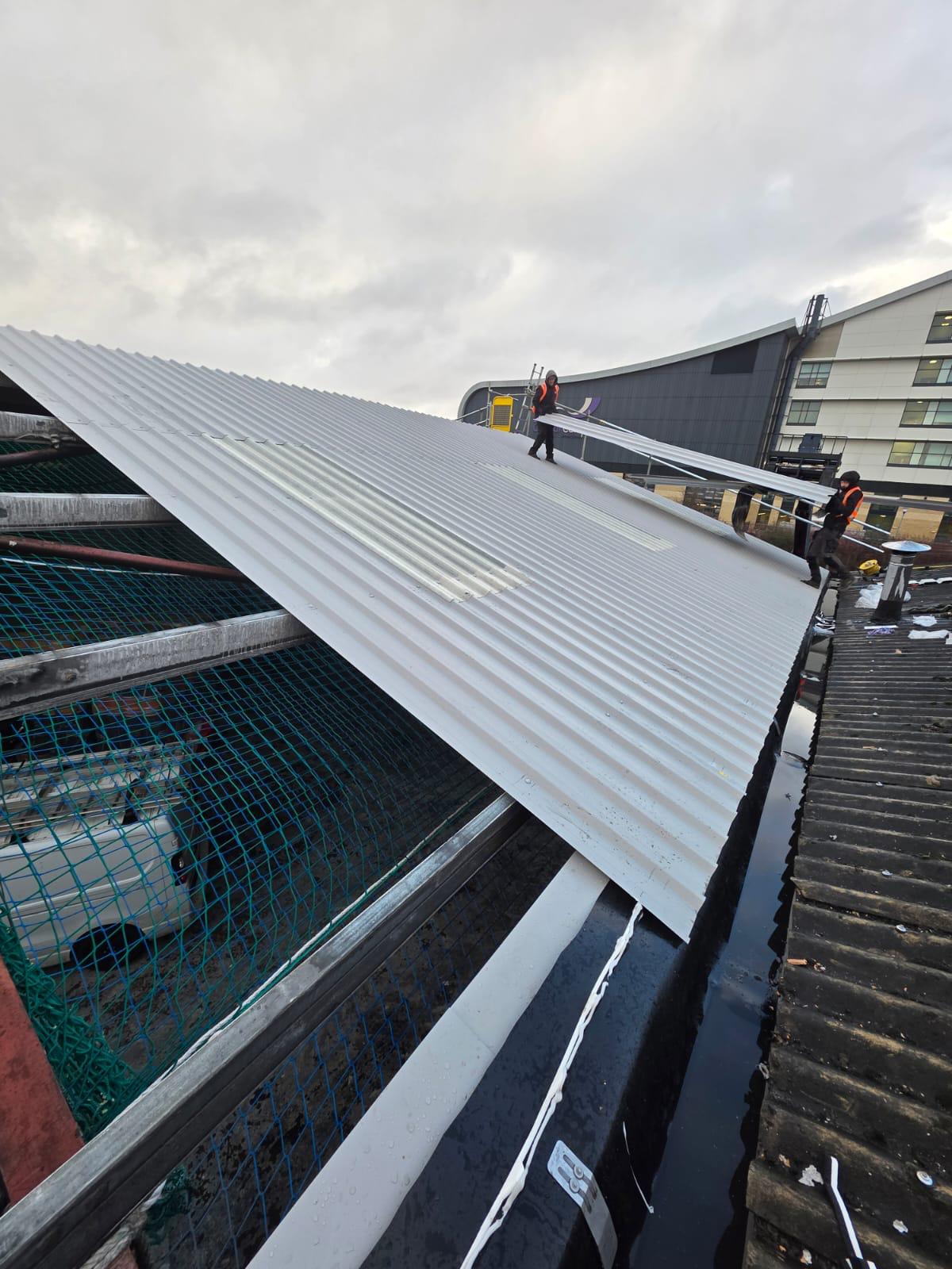 WhatsApp Image 2026-02-17 at 19.57.48 (1) Two roofing operatives manoeuvring a large profiled metal roof sheet into position during installation, with safety netting visible below and scaffolding to the side