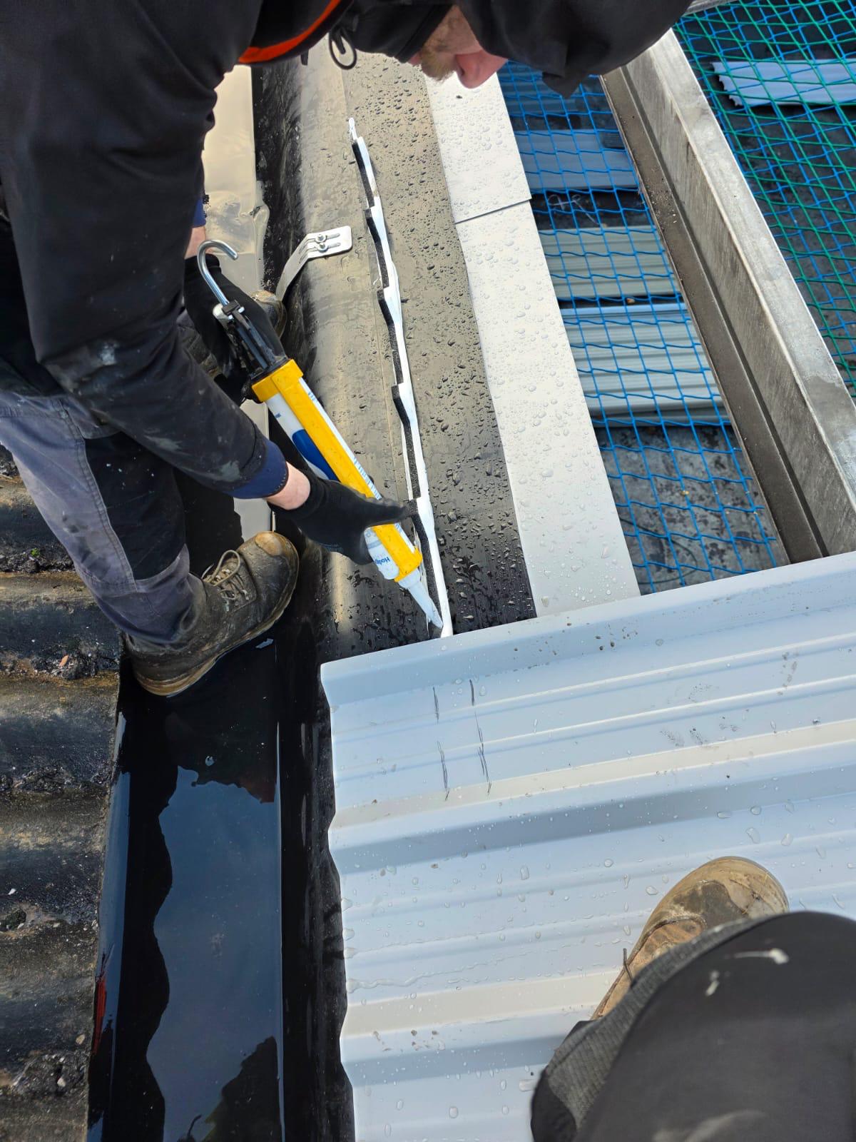 WhatsApp Image 2026-02-17 at 19.57.48 (3) Roofing operative applying sealant to a flashing junction between a new profiled metal roof sheet and a black gutterline, with safety netting visible in the background