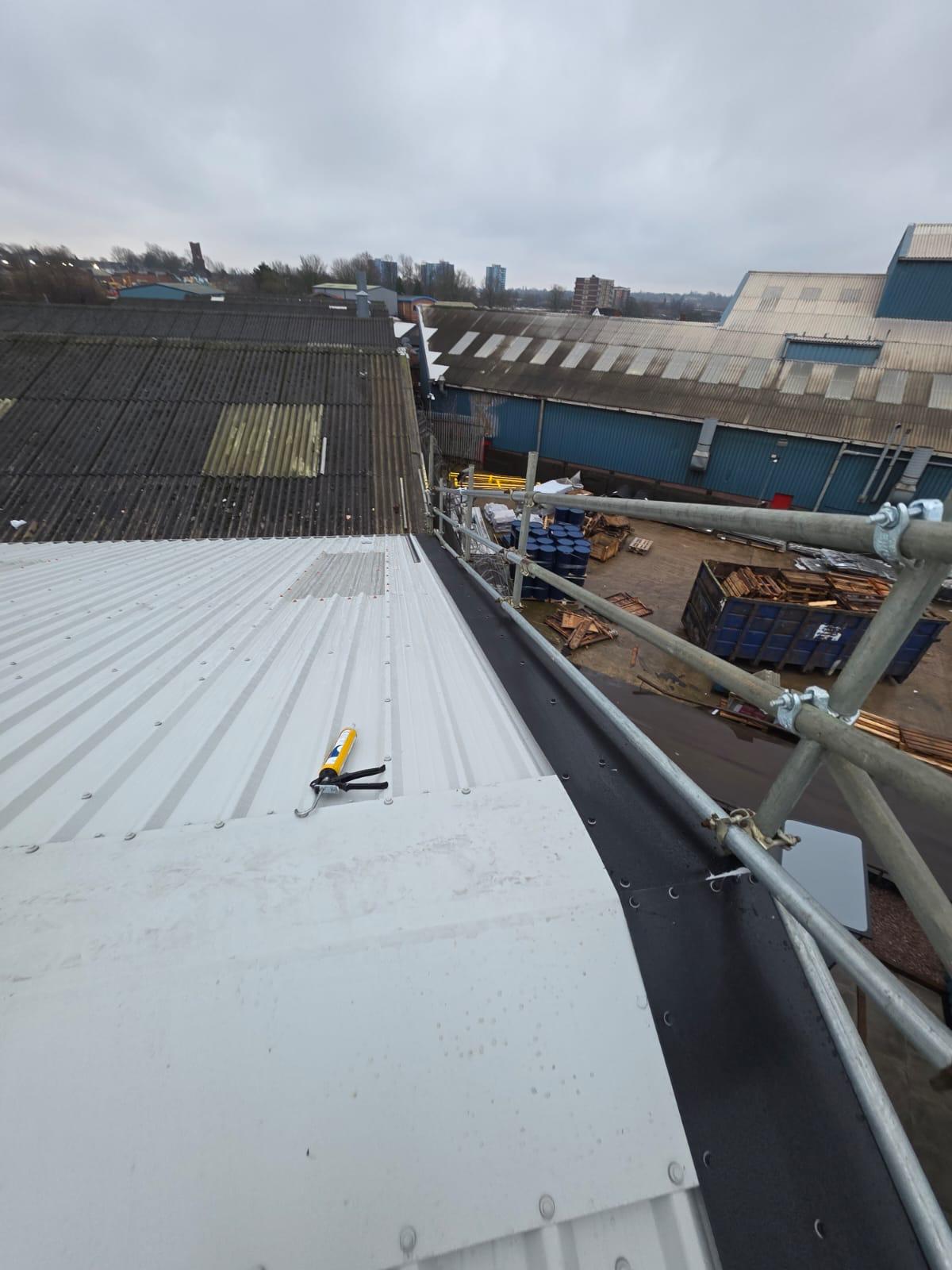 WhatsApp Image 2026-02-17 at 19.57.48 (5) New white profiled metal roof eaves detail showing a black gutterline and perimeter scaffolding, with a sealant gun resting on the roof surface and deteriorated asbestos cement roofs on neighbouring buildings visible in the background