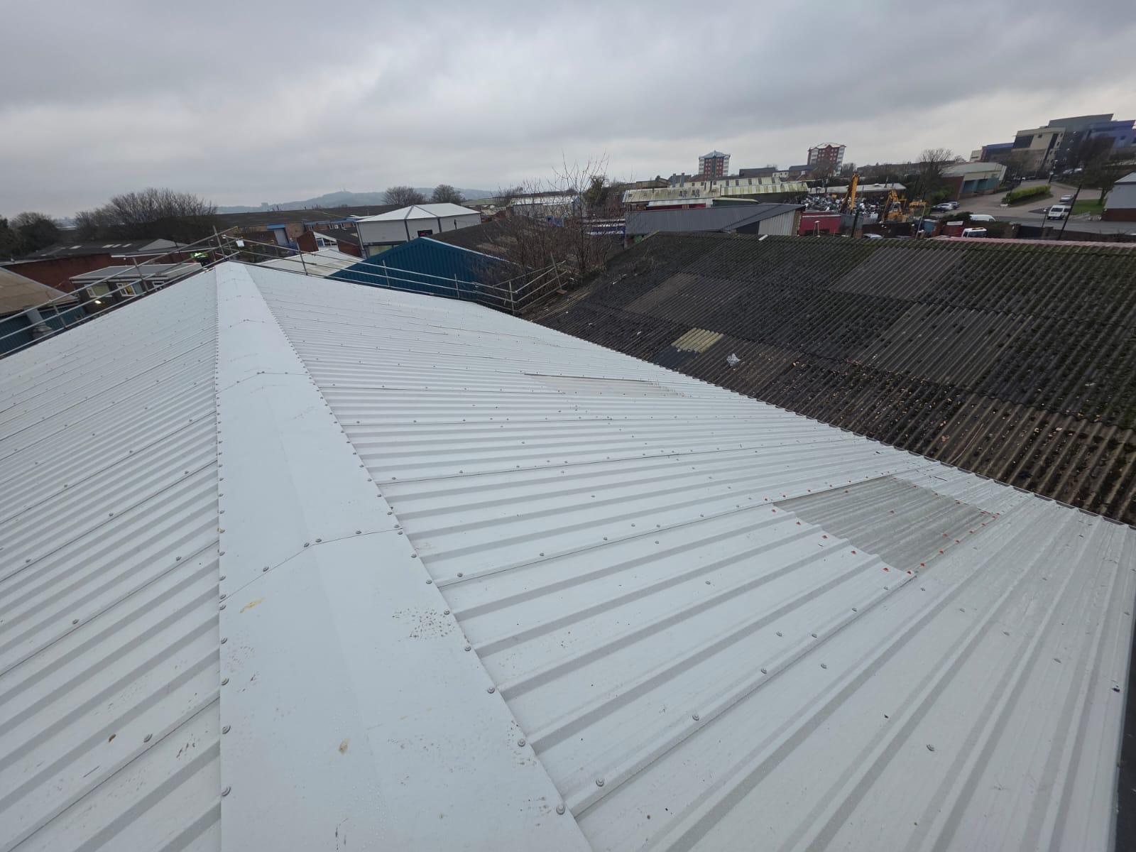 WhatsApp Image 2026-02-17 at 19.57.48 (6) Completed white profiled metal roof showing apex ridge and valley detail where two roof pitches meet, with deteriorated asbestos cement roofs on neighbouring commercial buildings visible beyond