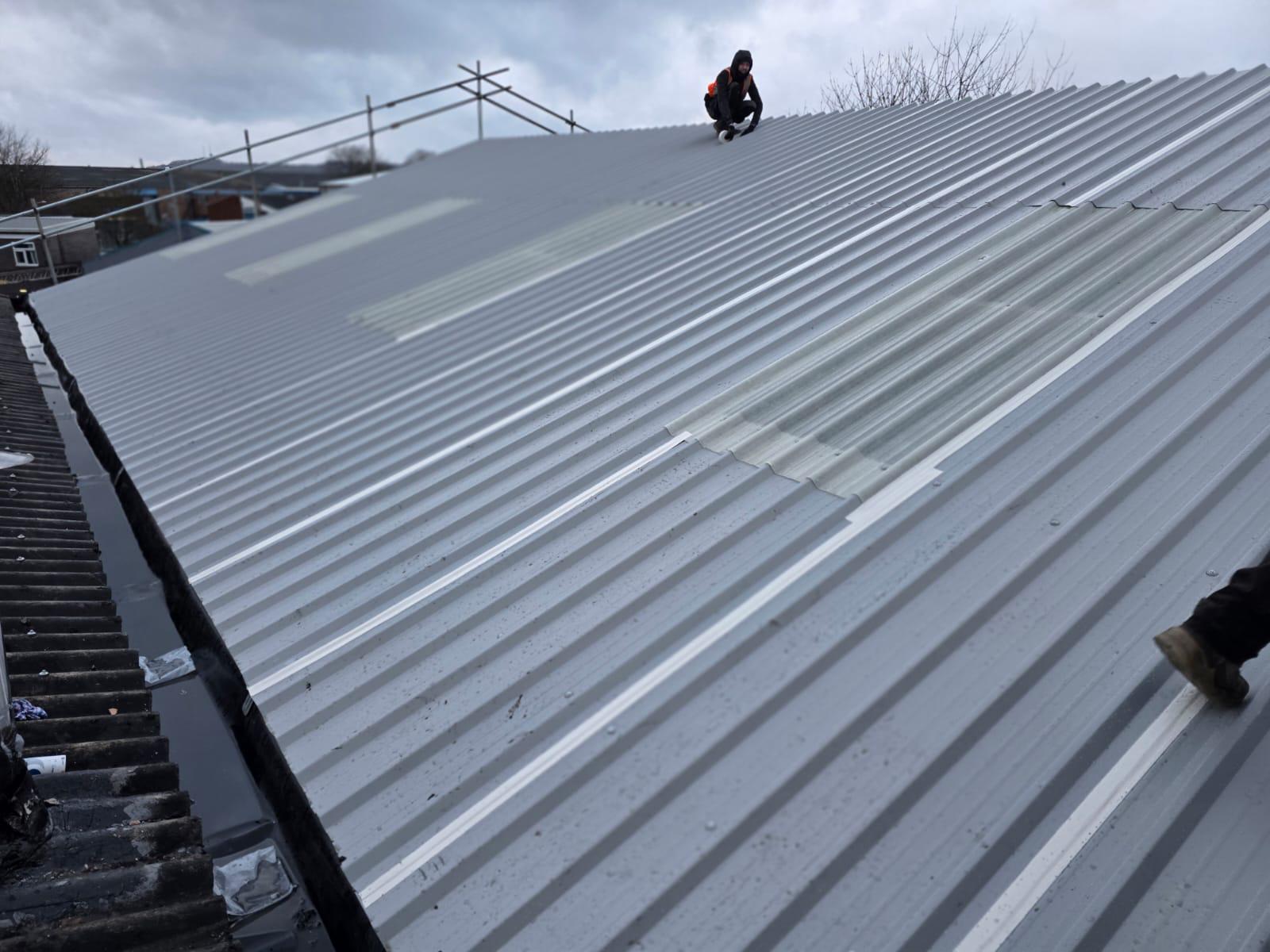 WhatsApp Image 2026-02-17 at 19.57.48 Roofing operative inspecting newly installed grey profiled metal roof sheets with polycarbonate rooflights, alongside a deteriorated asbestos cement roof on an adjacent building