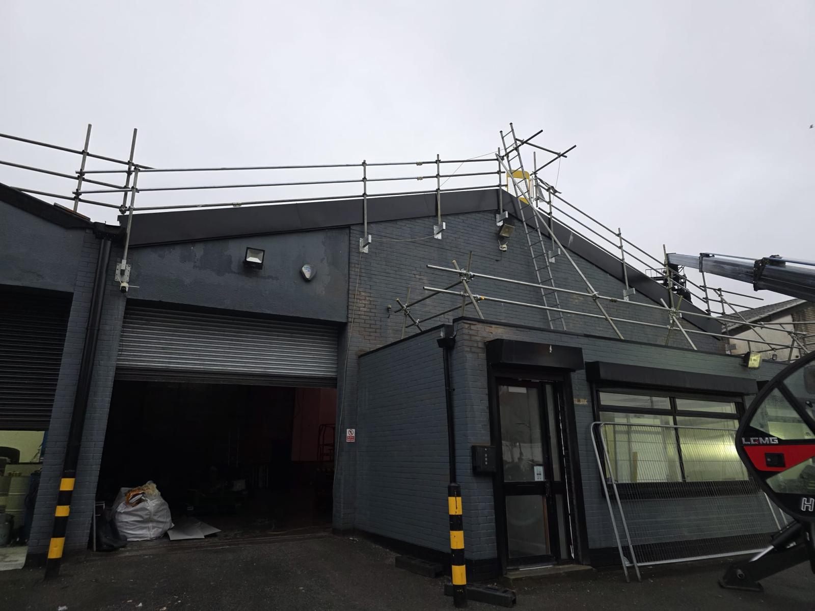 WhatsApp Image 2026-02-17 at 19.57.49 Front exterior of a commercial garage building with scaffolding and a telehandler in position ahead of roof replacement works, showing roller shutter doors and a pitched roof gable end