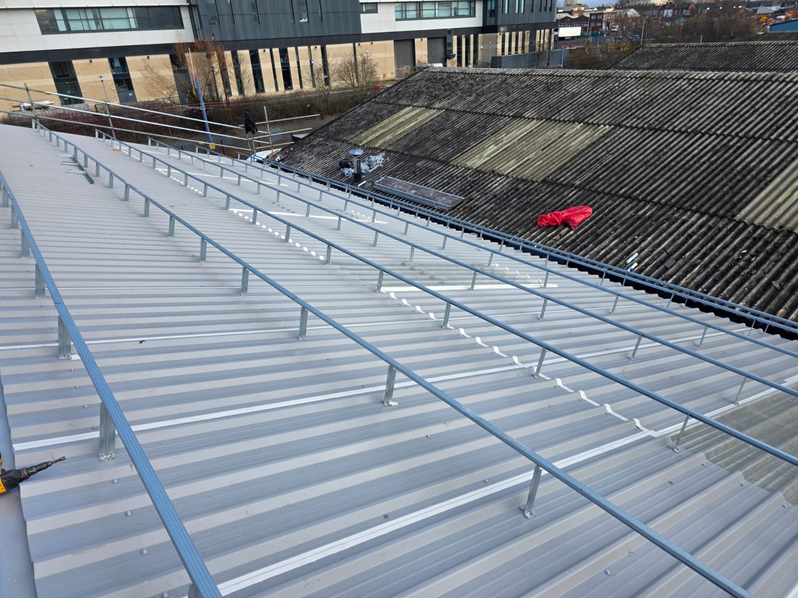 Newly installed grey profiled metal roof sheeting with steel safety handrails, with a deteriorated asbestos cement roof visible on the adjacent building