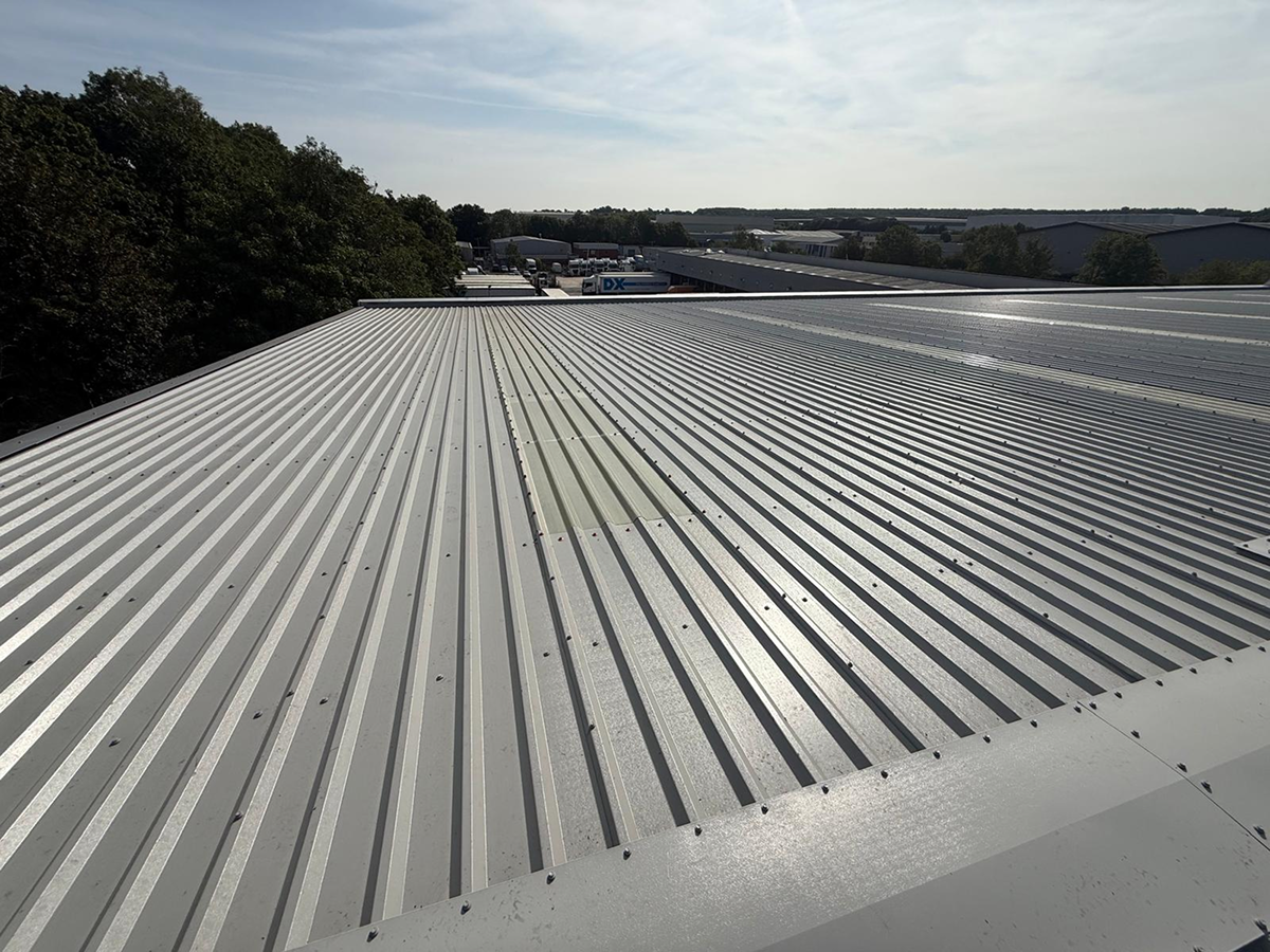 Completed grey profiled metal roof stretching across to the eaves, with neighbouring industrial units and trees beyond