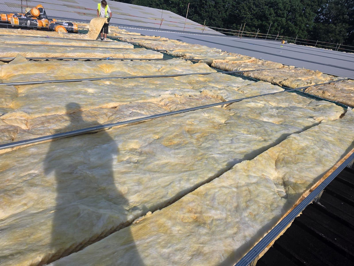 Roofing operative laying quilt insulation between spacer bars over an existing dark industrial roof, with new roof sheets installed alongside