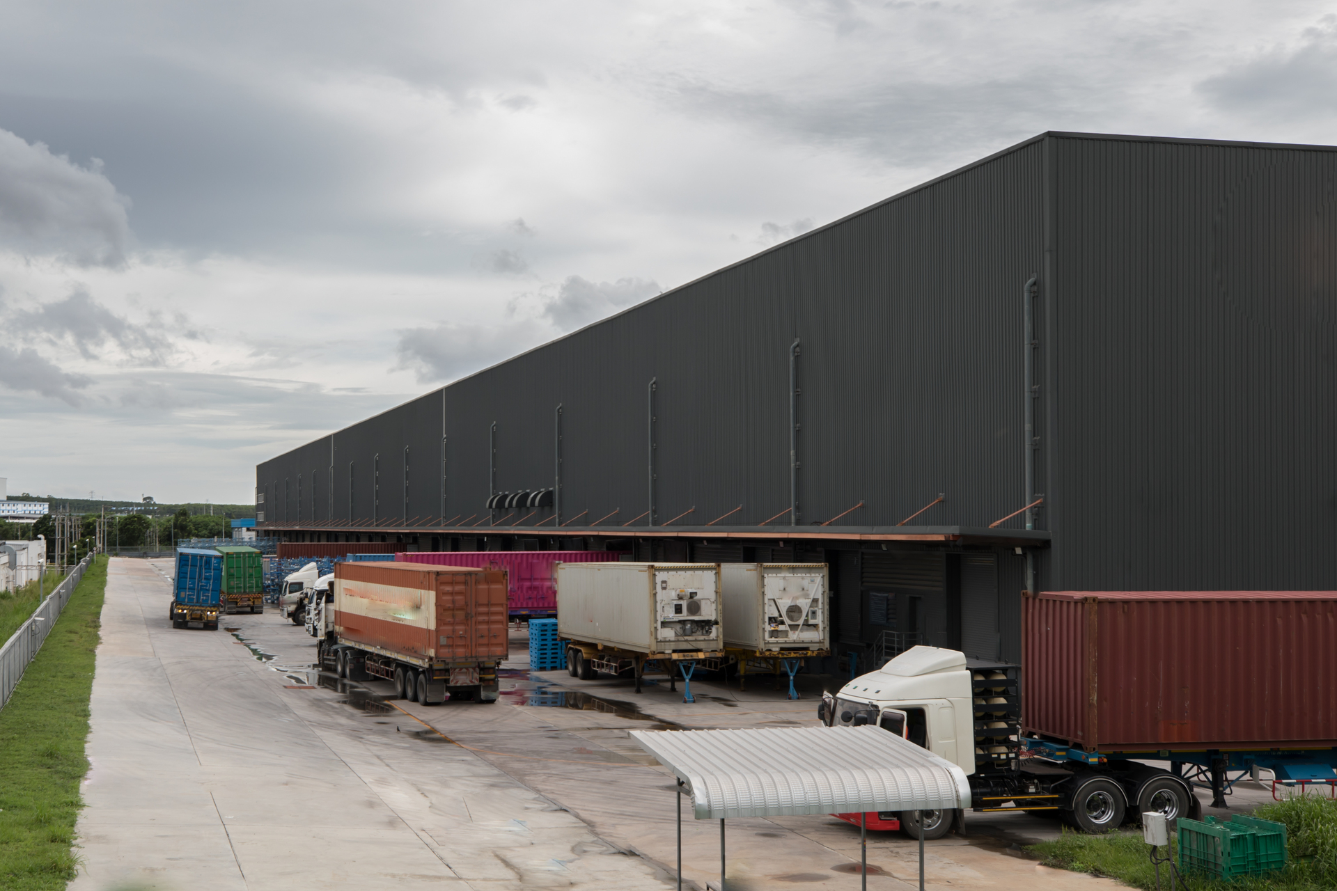 Trucks with shipping containers parked at warehouse loading docks under a cloudy sky.